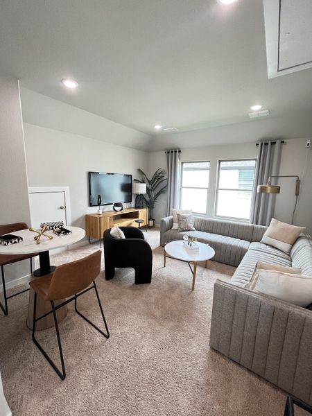 A cozy living space with modern furniture, neutral tones, plush carpet, and natural light filtering through wide windows.