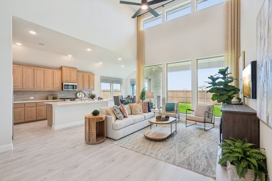 Open-concept living room with two-story ceiling, ceiling fan, and kitchen visible in background with light wood cabinets Open-concept living room with two-story ceiling, ceiling fan, and kitchen visible in background with light wood cabinets