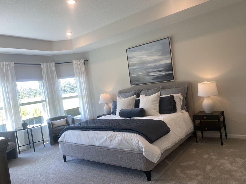 A serene bedroom with cozy gray carpeting, plush bedding, and elegant lighting, complemented by large windows and soft curtains.