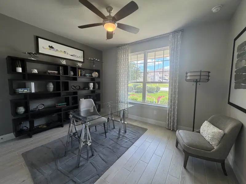A modern office with sleek glass desk, stylish gray shelving, and large window, complemented by chic decor and a cozy chair.