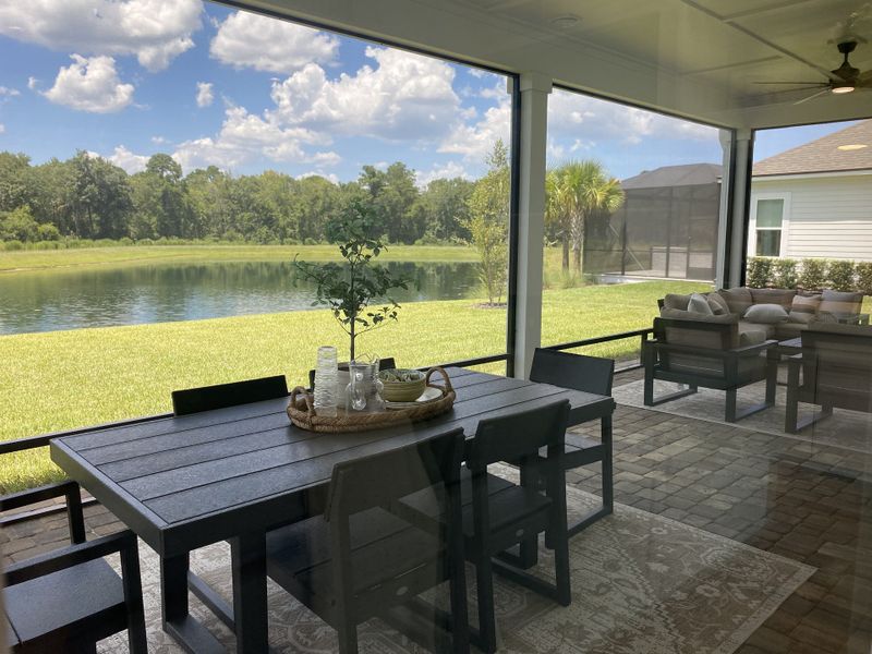 A serene patio with elegant outdoor furniture overlooking a tranquil pond and lush greenery under a bright blue sky.