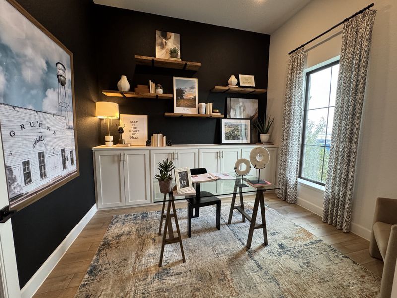 A cozy home office with dark accent wall, modern artwork, white cabinets, and large window. Stylish decor enhances warmth.
