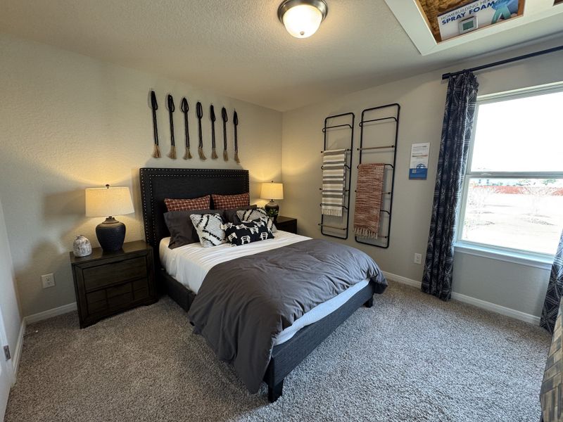 A cozy bedroom with a plush bed, elegant lamps, and tasteful wall decor. Carpeted floor and a window with patterned curtains.