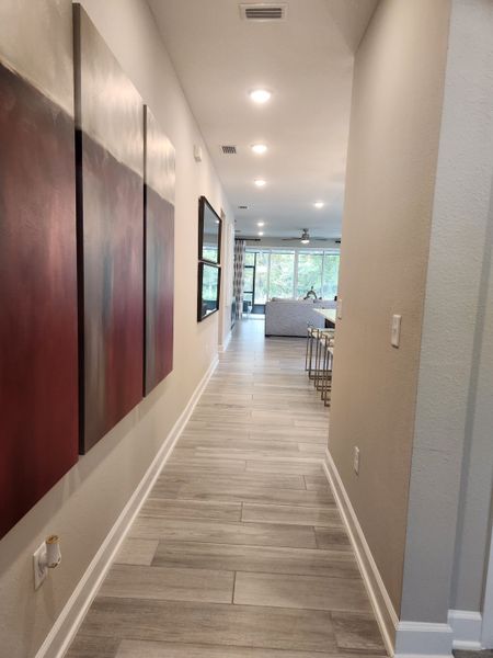 A modern hallway with elegant wood-look tile flooring, contemporary artwork, and recessed lighting leading to a bright living area.