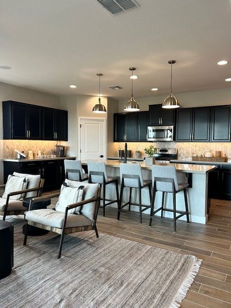 A modern kitchen with sleek black cabinets, stainless steel appliances, and cozy seating, featuring elegant pendant lighting.
