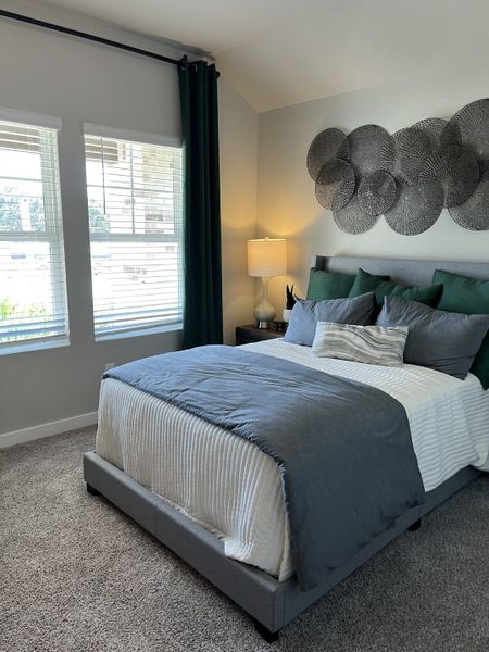 A cozy bedroom with a plush bed, teal accents, textured art, and soft carpet, complemented by large windows and elegant drapery.