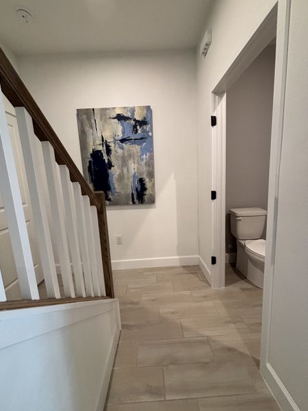 A modern hallway featuring sleek tile flooring, a wooden staircase, and abstract wall art adjacent to a compact bathroom.
