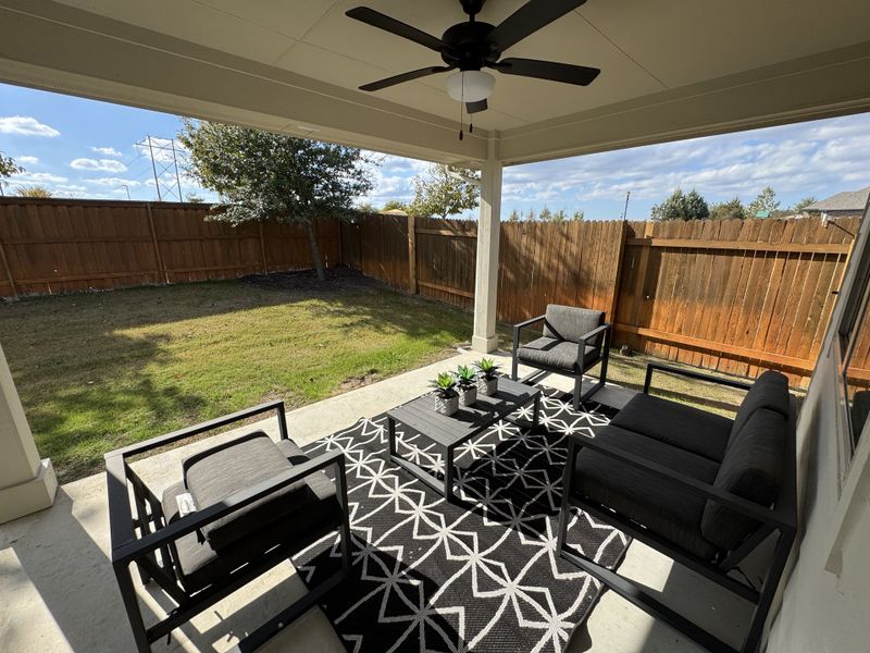A cozy patio with modern black furniture, patterned rug, and a ceiling fan overlooking a fenced yard.