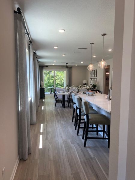 An elegant dining area features a long table, stylish pendant lighting, and large windows overlooking greenery.