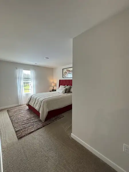 A cozy bedroom featuring a plush red bed, soft carpet, and ample natural light from a large window. A cozy bedroom featuring a plush red bed, soft carpet, and ample natural light from a large window.