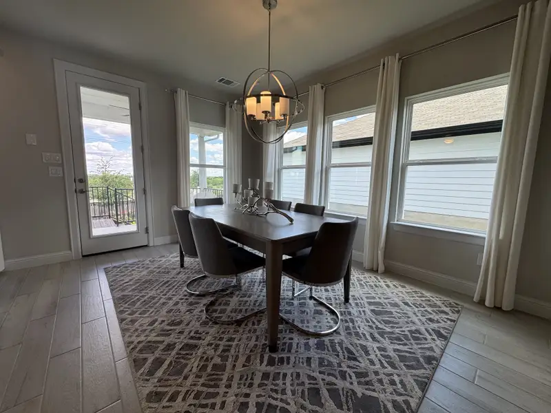 A bright dining room with large windows, sleek chandelier, and modern table on a patterned rug.