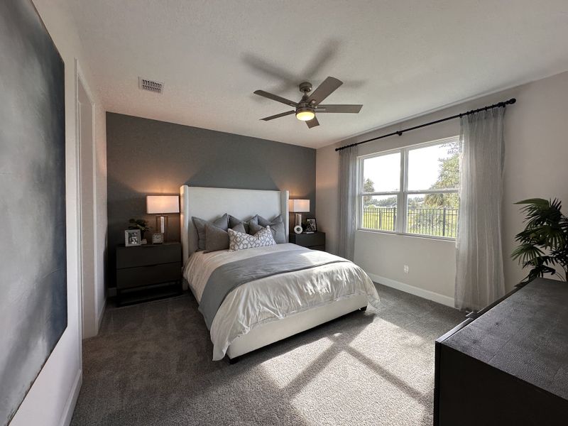 A cozy bedroom with a modern ceiling fan, soft gray accents, and warm lighting, featuring large windows overlooking greenery.
