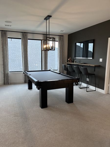 A modern game room featuring a sleek black pool table, cushioned bar stools, and elegant pendant lighting with large windows.