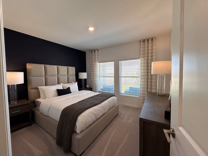 A cozy bedroom featuring a plush upholstered bed, dark accent wall, and soft lighting with large windows.