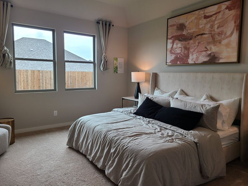 A cozy bedroom featuring a plush bed, abstract art, and soft natural light from large windows.