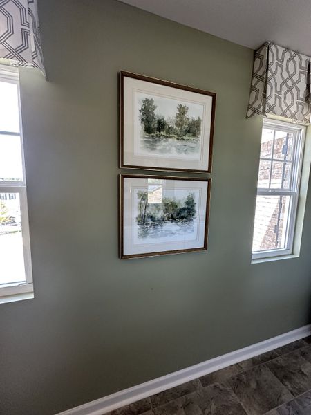 A cozy corner featuring framed landscape art, soft green walls, patterned valances, and abundant natural light through dual windows.