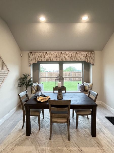 A cozy dining area with a dark wooden table, elegant chairs, and a window seat with pillows, opening to a lush garden view.