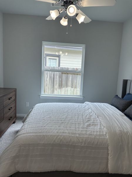 A cozy bedroom featuring a plush bed, wooden dresser, and bright window lighting under a decorative ceiling fan. A cozy bedroom featuring a plush bed, wooden dresser, and bright window lighting under a decorative ceiling fan.