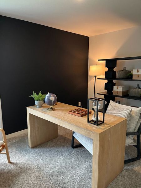 A modern office with a sleek wood desk, black accent wall, stylish shelving, and cozy seating.