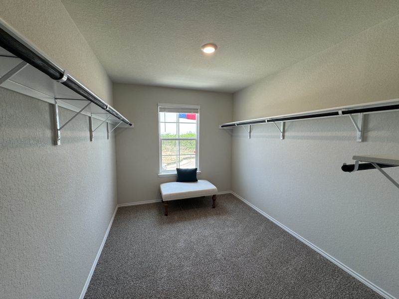 A spacious walk-in closet with two parallel hanging rails, a bench under a window, and soft carpet flooring.