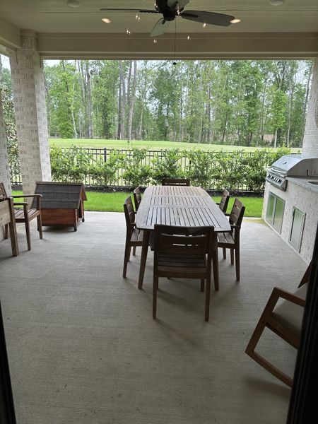 A serene covered patio with a wooden dining set, ceiling fan, and built-in grill overlooking lush greenery.