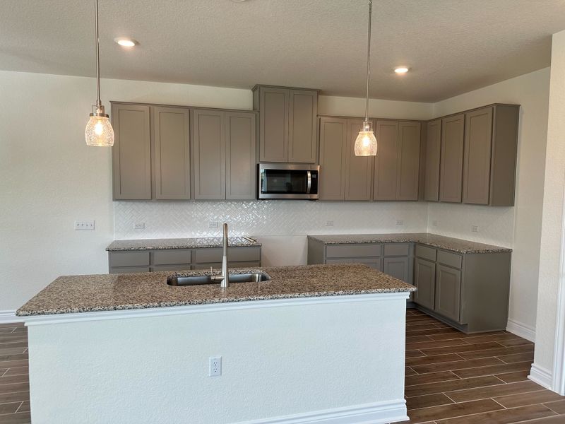A modern kitchen with granite countertops, sleek gray cabinets, pendant lighting, and wood-look tile flooring.