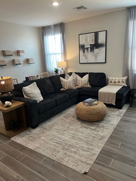 A cozy living room with a dark sectional, abstract art, and a woven ottoman on a textured rug.