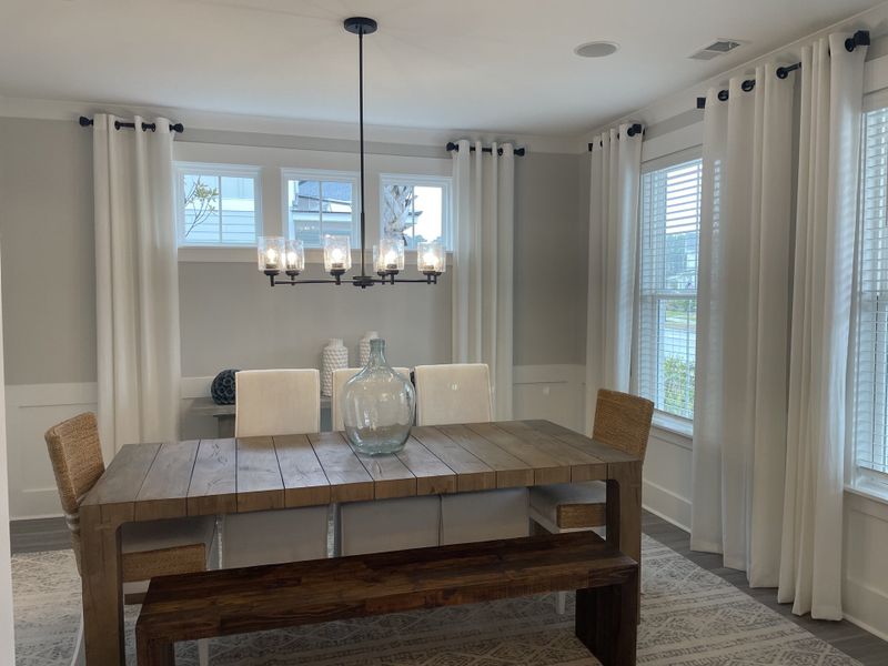 A charming dining area with a rustic wooden table, woven chairs, and a stylish chandelier, surrounded by bright windows and soft white curtains.