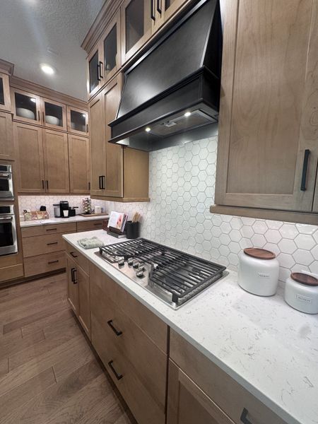 A modern kitchen featuring wood cabinets, a sleek black hood, and hexagonal tile backsplash with quartz countertops.