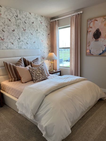 A cozy bedroom with floral wallpaper, plush bedding, pink curtains, and serene artwork.