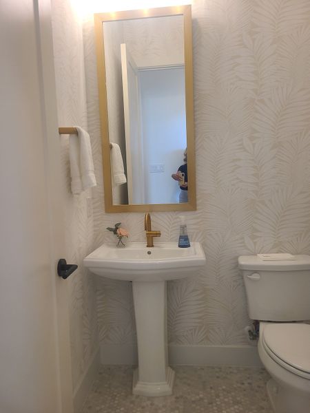 A chic bathroom with a gold-framed mirror, pedestal sink, and light leaf-patterned wallpaper for a stylish touch.