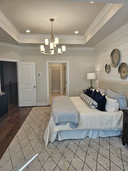 A cozy bedroom with elegant tray ceiling, modern chandelier, plush bedding, and tasteful décor on a soft patterned rug.