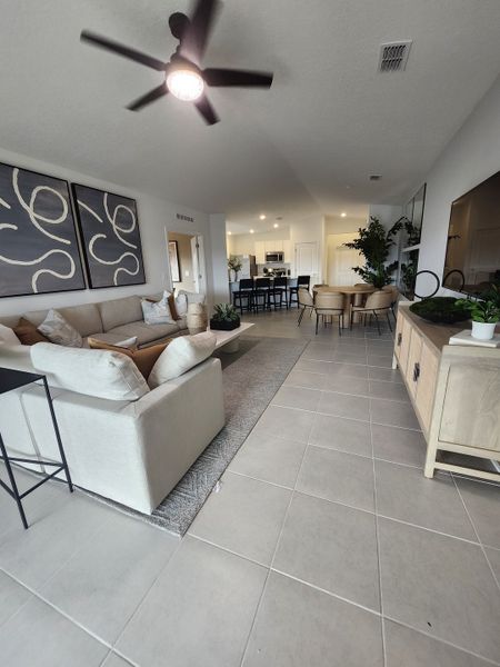 A modern living room with a beige sofa, tile floors, abstract art, and open dining area under sleek ceiling fan lighting.