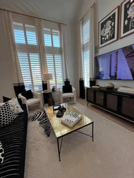 A chic living room with tall windows, elegant drapes, modern decor, and a gold-accented coffee table.