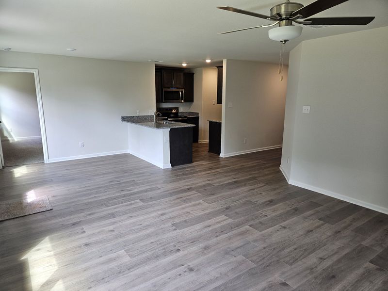 A modern living space featuring wood-style flooring, an open kitchen with dark cabinets, and a ceiling fan.