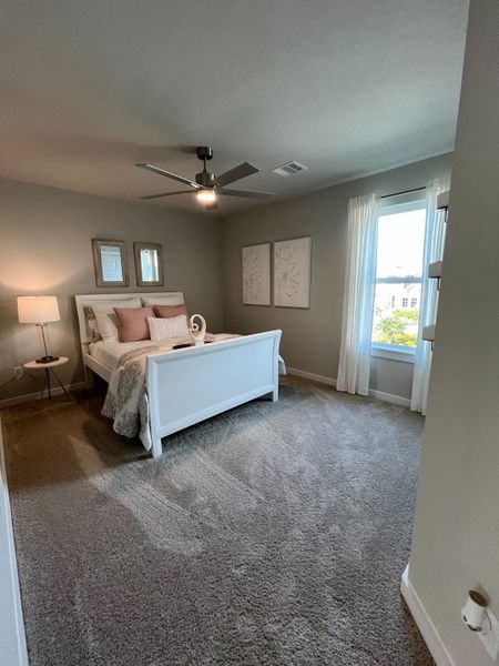 A cozy bedroom featuring a white bed, soft gray carpet, modern ceiling fan, and gentle natural light through sheer curtains. A cozy bedroom featuring a white bed, soft gray carpet, modern ceiling fan, and gentle natural light through sheer curtains.