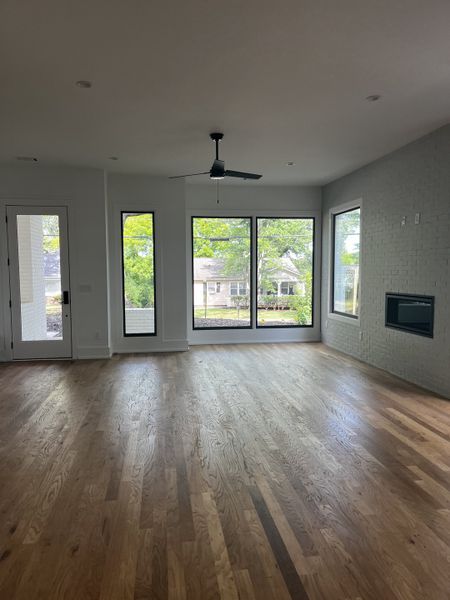 A spacious living room with large windows, hardwood floors, modern ceiling fan, and a built-in fireplace.
