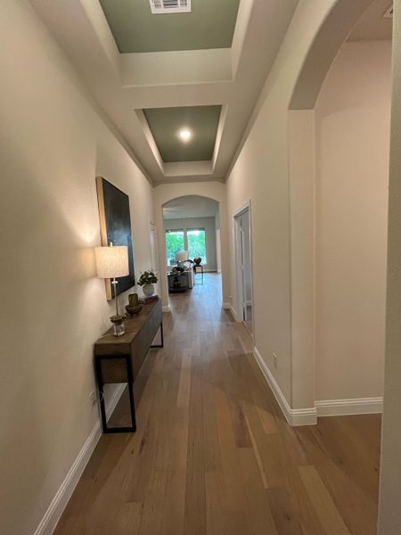 A bright hallway with wooden floors, tray ceiling, and elegant console table leading to a light-filled living area.