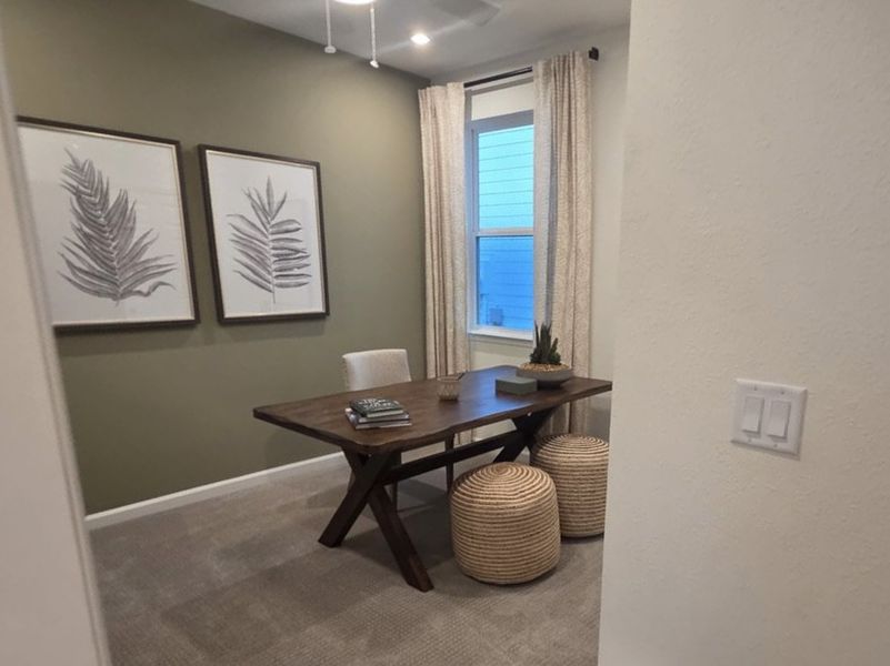 A cozy home office with a wooden desk, woven stools, green accent wall, and botanical art.