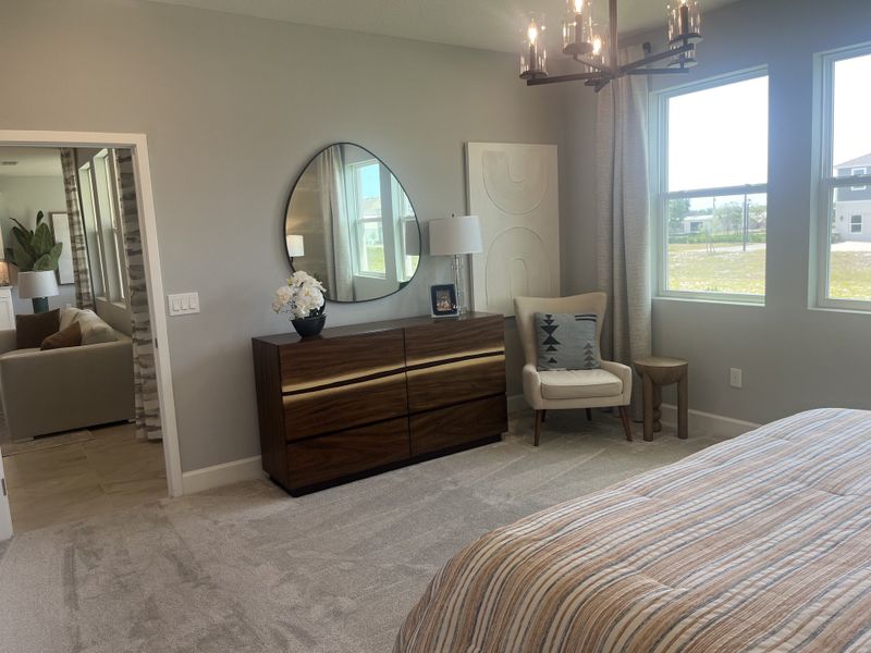 A cozy bedroom with a modern wood dresser, chic mirror, and plush chair. Natural light floods through large windows.