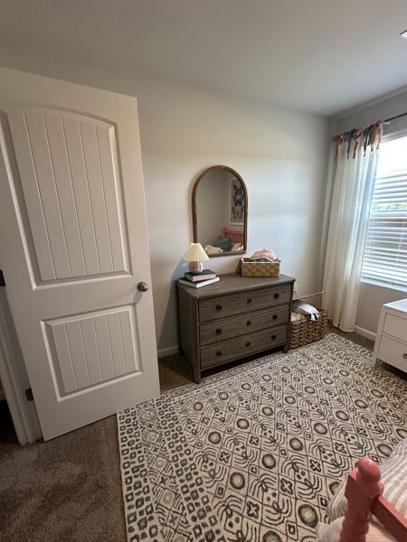 A cozy bedroom with a patterned rug, wooden dresser, elegant mirror, and soft lighting, creating a warm and inviting atmosphere.