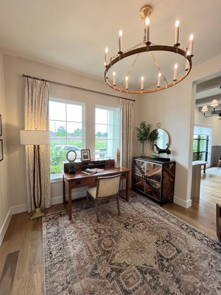 A cozy study with a wooden desk, elegant chandelier, and large windows overlooking a lush garden.