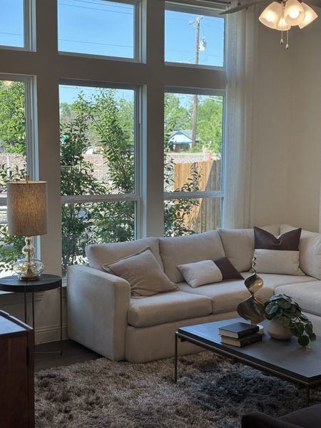A cozy living room with a beige sofa, plush rug, and large windows offering natural light and outdoor greenery views.