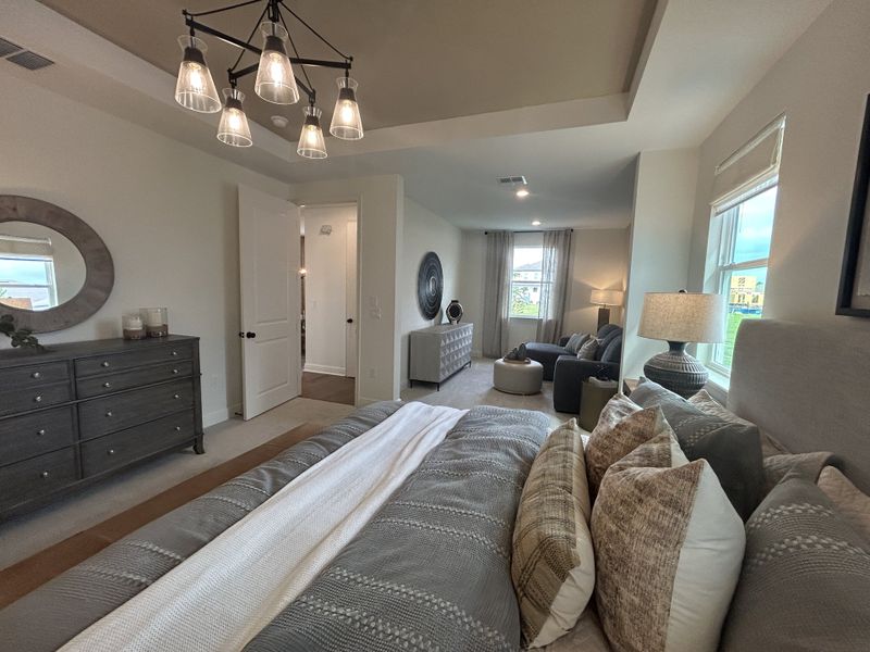 A cozy bedroom with elegant bedding, a modern chandelier, and a seating area, bathed in natural light.