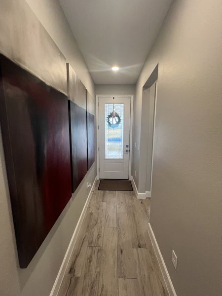 A modern hallway with gray walls, wood-look flooring, and abstract wall art leading to a glass-paneled door adorned with a wreath.