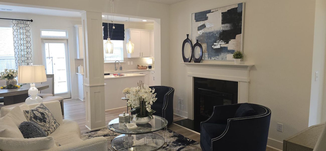 A cozy living room with a modern fireplace, elegant decor, and an open view into a bright kitchen with pendant lighting.