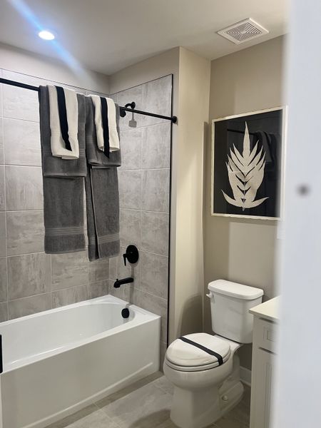 A modern bathroom featuring a tiled bath, sleek black fixtures, and minimalist decor with a leafy wall art piece.