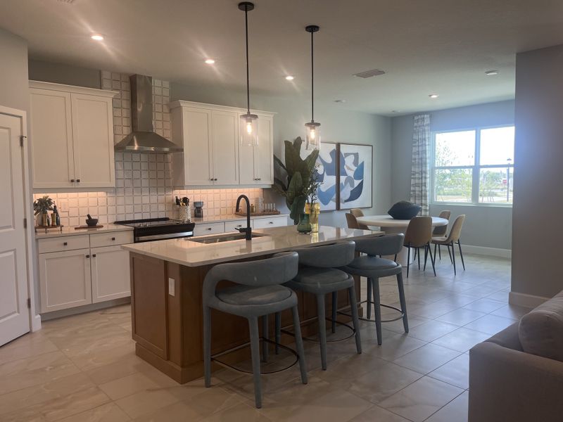 A modern kitchen with white cabinets, tiled backsplash, island seating, and a cozy dining area with large windows.