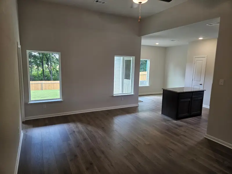 An open, airy room with wood floors, large windows, and a central island, offering a view of the lush backyard.