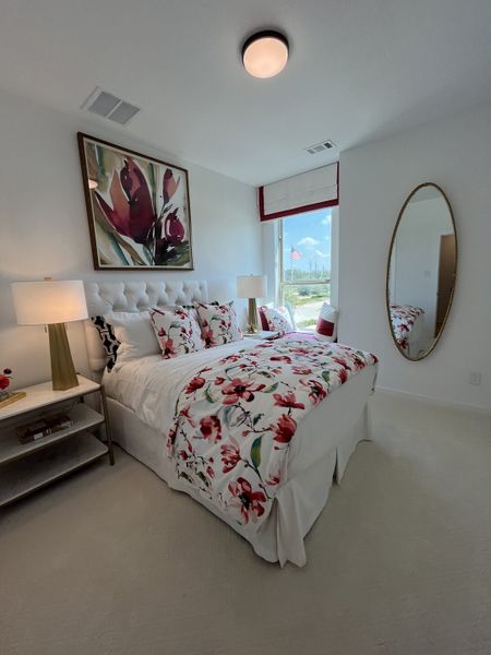 A bright bedroom with floral bedding, elegant artwork, and a large oval mirror enhancing the airy, modern design.